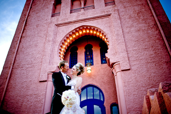the happy couple embracing as they stand in front of a beautiful dark pink stucco church with purple door and window frames - photo by New Mexico based wedding photographers Twin Lens